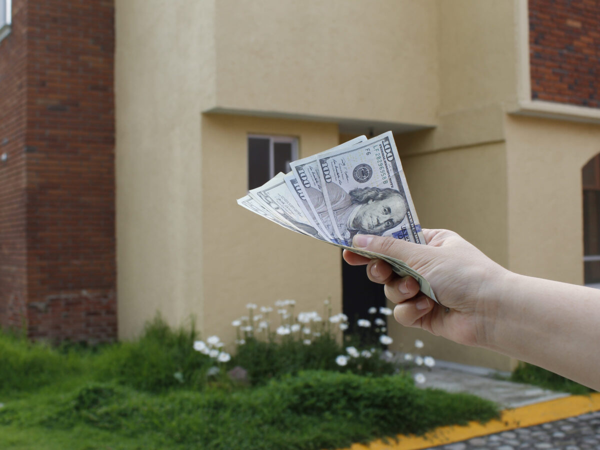Remote Workers Are Learning the Hard Way That the Wrong State Can Shrink a Good Paycheck 4 hand of a woman holding american banknotes and background with facade of a house