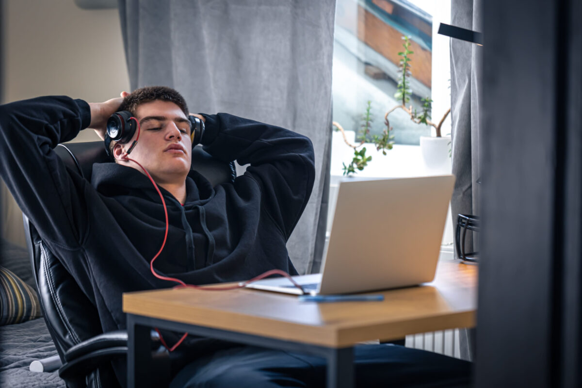 A tired teenager guy in headphones sits in front of a laptop in his room