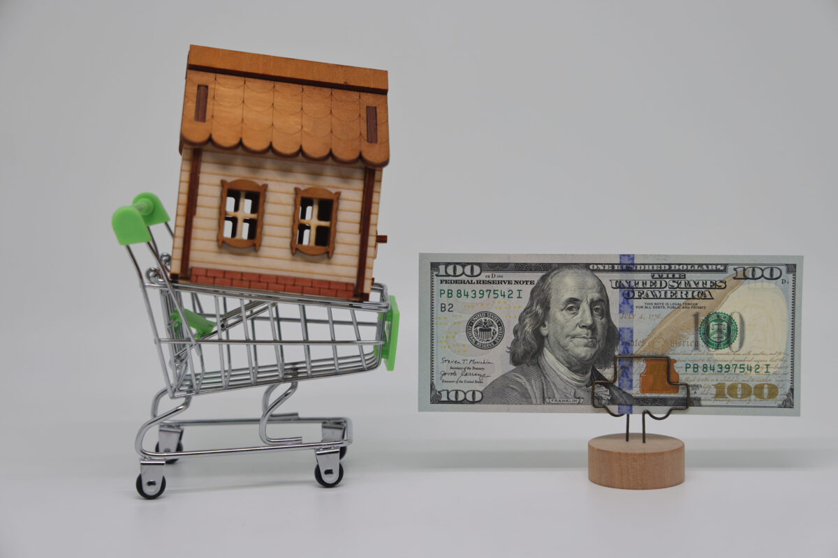 A toy grocery cart and a toy house stand next to a banknote on a stand, real estate purchase and mortgage