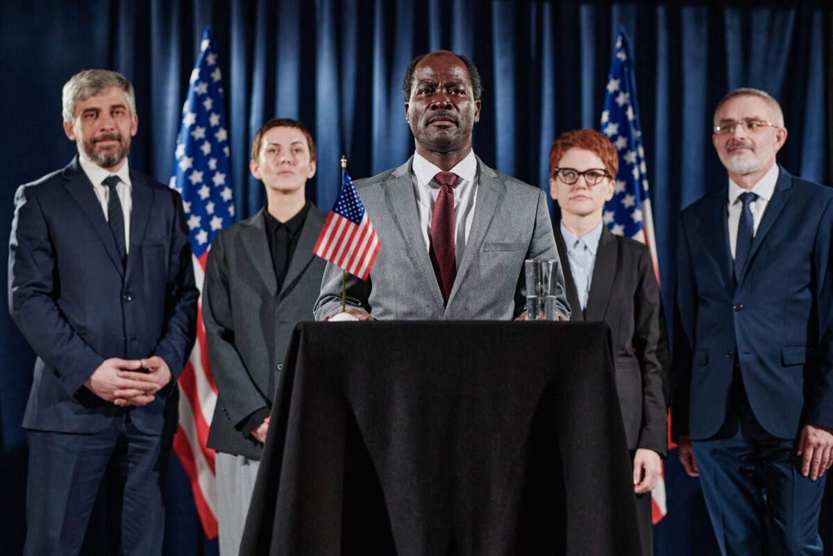 Portrait of African mature leader standing at tribune at press conference with his business team behind him