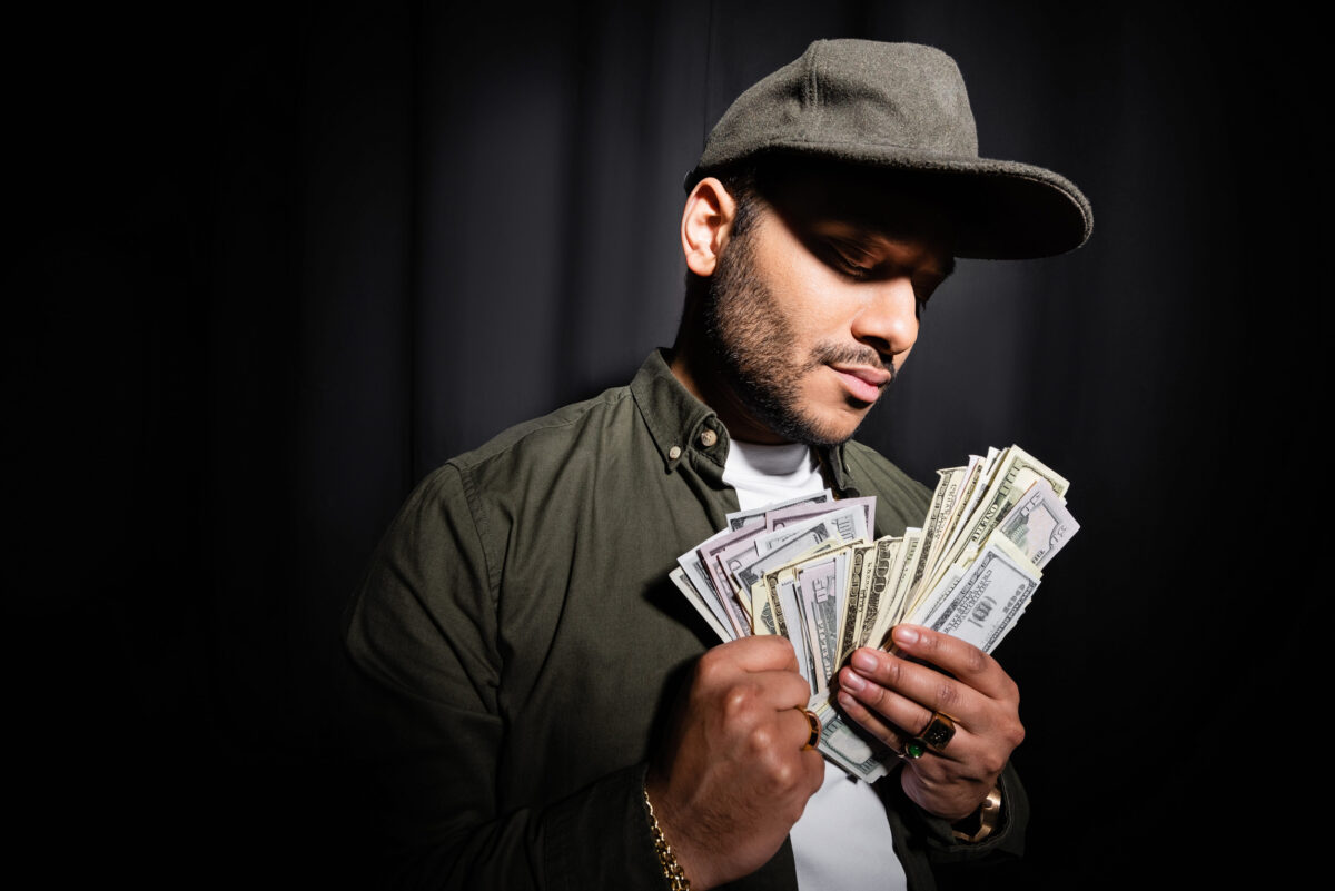 rich indian hip hop performer in cap holding dollar banknotes on black