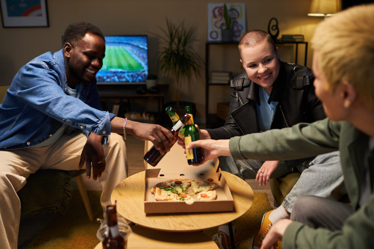 10 Cheap Meals Americans Crave Most When They’re Financially Burned Out 4 Medium full shot of three diverse young adults toasting bottles of beer while watching football and eating pizza at home