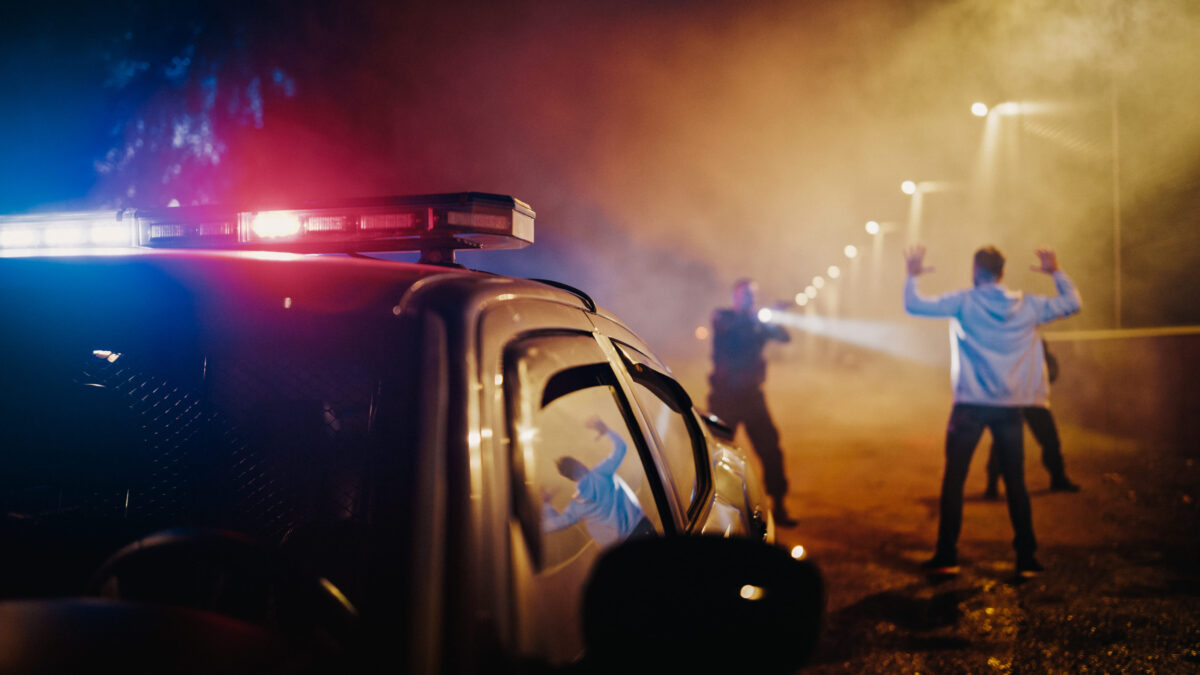 Two Police Officers Aim at a Fellon. Cops Using a Weapon to intimidate the Suspect. Fellon Raising Hands in Compliance. Law Enforcement Arresting a Dangerous Man. Camera Focus on Police Car Siren