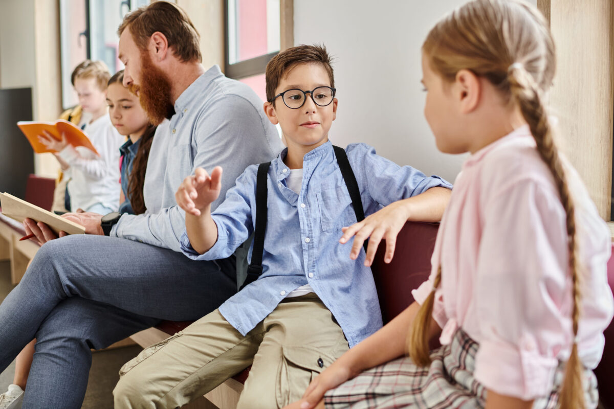 A male teacher instructs a group of diverse kids in a bright, lively classroom setting, sitting close to each other and engrossed in learning.