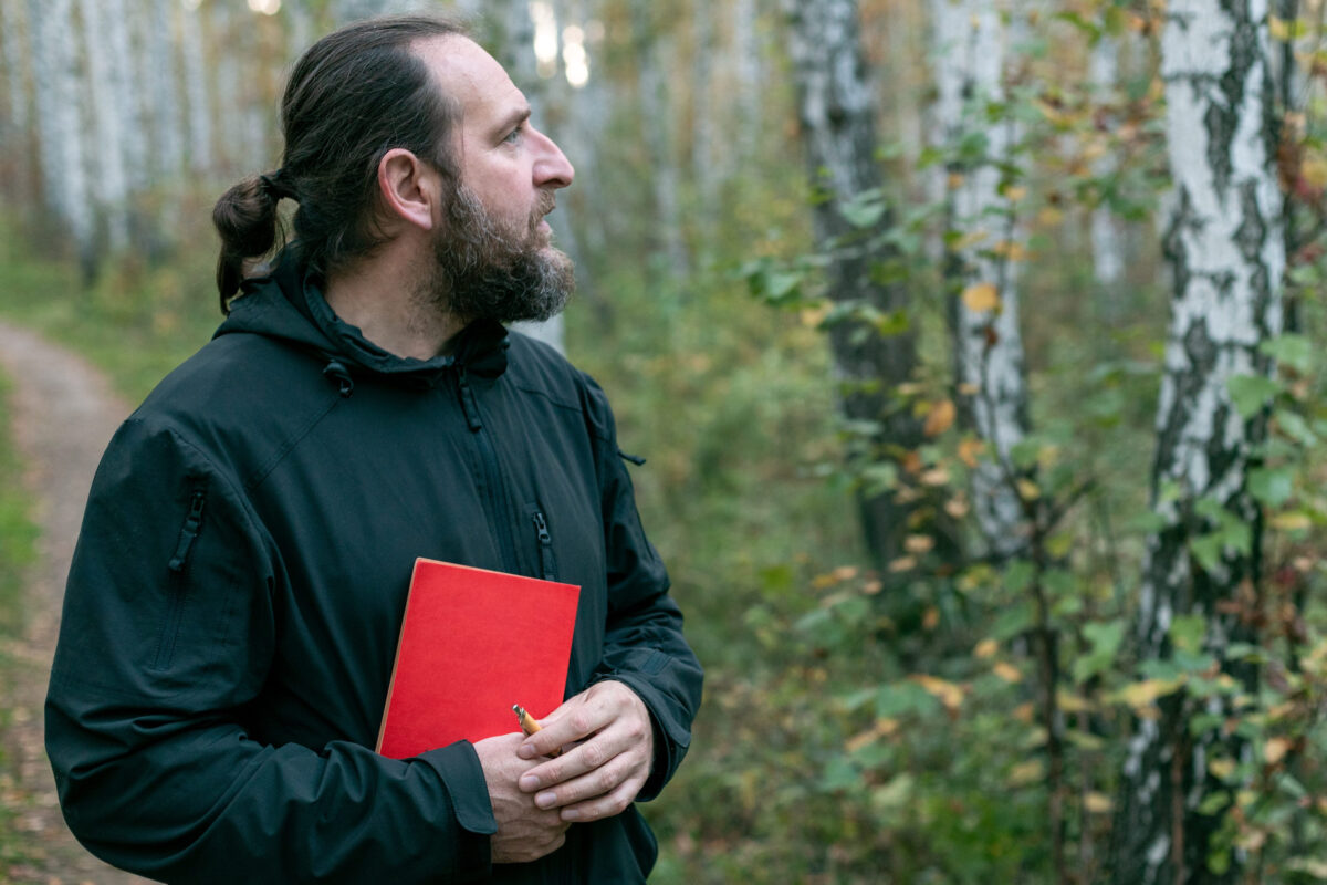 10 Spiritual Trends That Are More Dangerous Than They Seem 1 A man with a beard and long hair holds a red book in his hands. The scene is calm and peaceful, as the man is surrounded by nature.