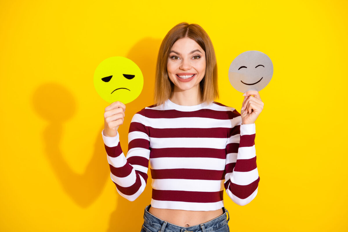 Attractive young woman holding two illustrated emotion signs against vibrant yellow background