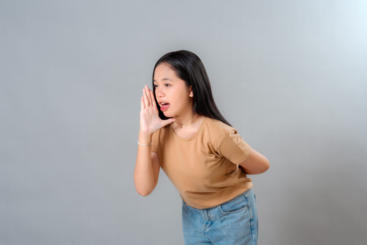 Portrait of a young Asian woman speaking with hand near her mouth, expressing announcement or gossip, showing open communication and confident expression, on a plain grey background.