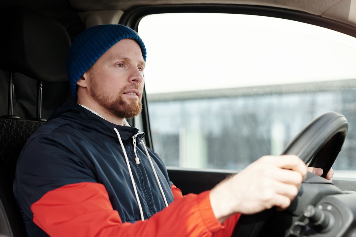 A bearded man wearing a hoodie and knitted cap driving a car, focused and attentive.