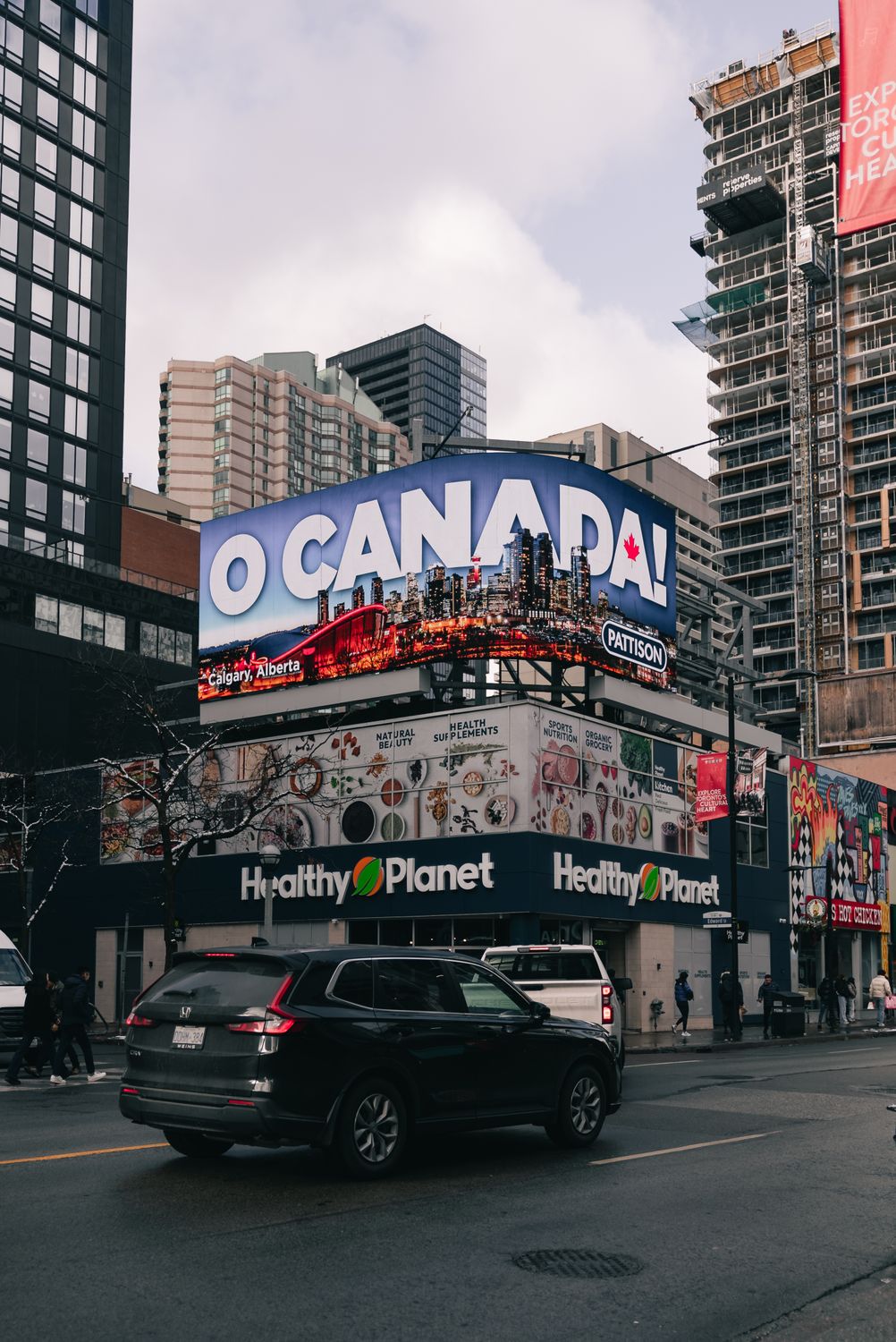 Everyone Wants to Move to Canada… But the Reality Is Not What You Expect 1 A bustling street in Toronto with a prominent 'O Canada' billboard, cars, and skyscrapers.
