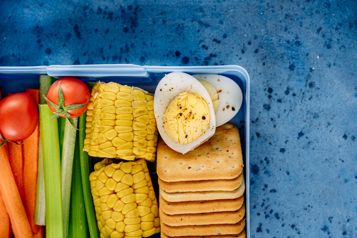 A colorful lunchbox featuring boiled eggs, corn, crackers, and fresh vegetables on a blue background.