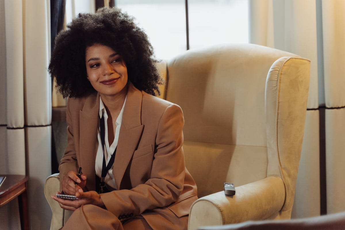 A confident woman in a brown blazer sits in an interview setting, taking notes.