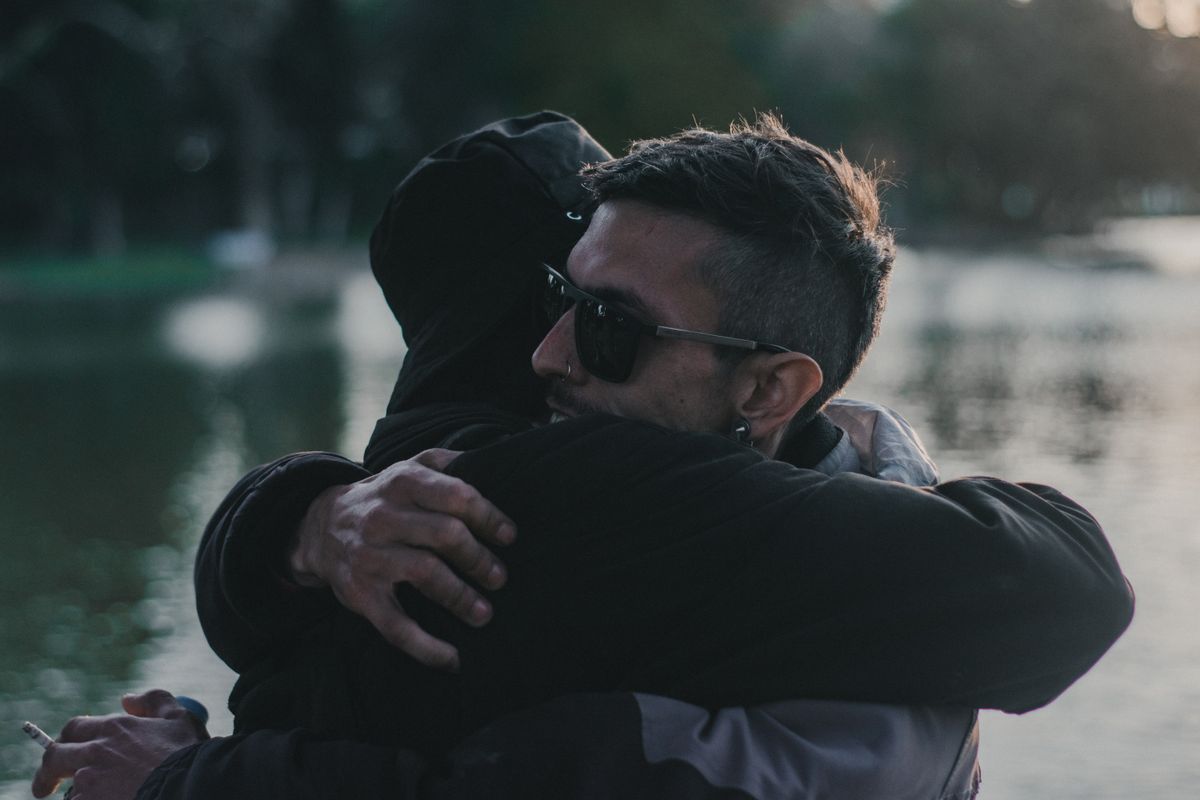 A heartfelt embrace between two adults near a calm lakeside during daylight.