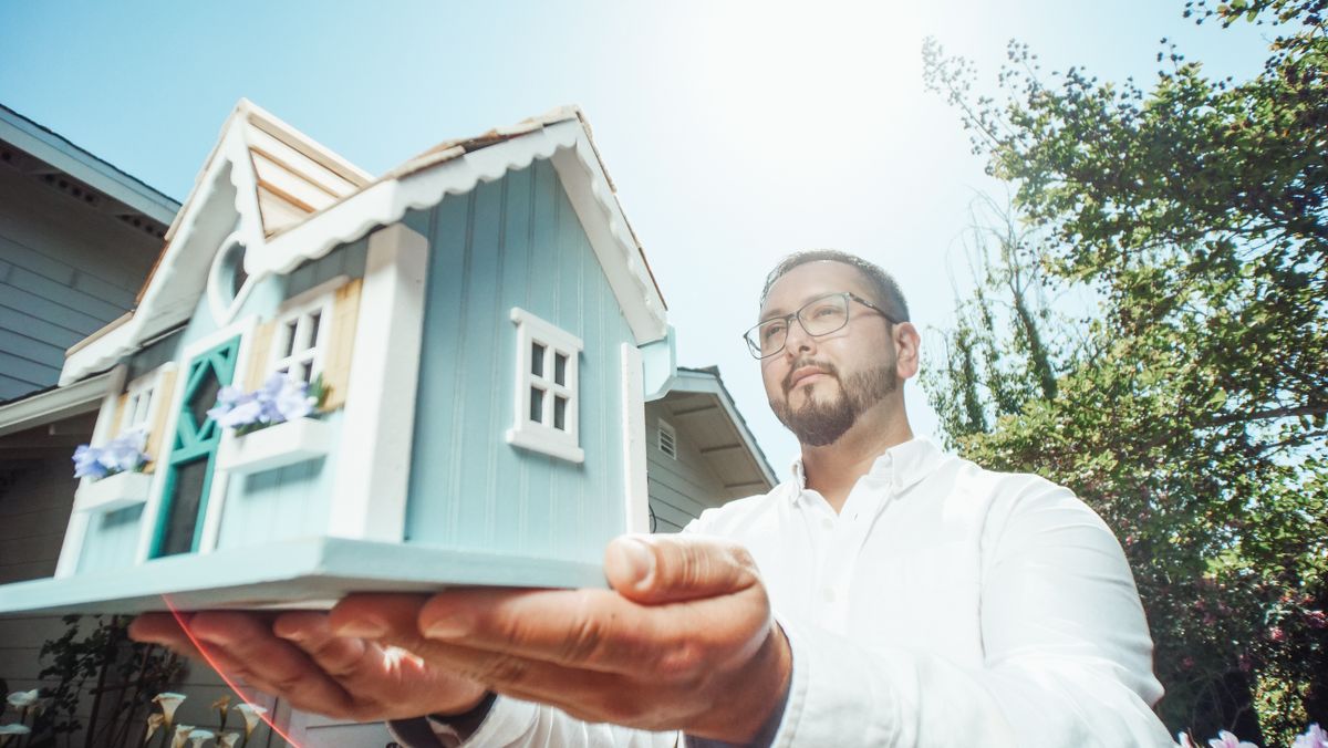A man holds a miniature house model outdoors on a sunny day, symbolizing home buying.