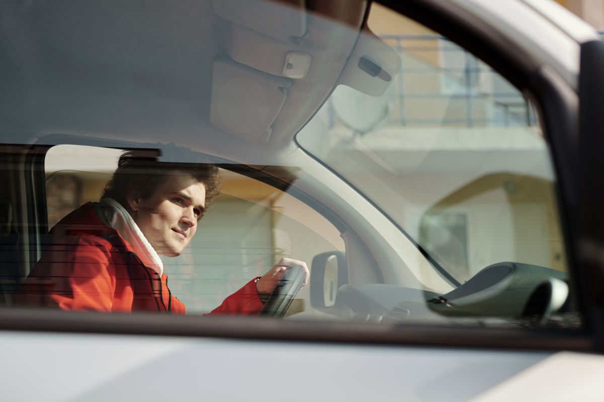 A man in a red jacket driving a car, seen through the window on a sunny day.