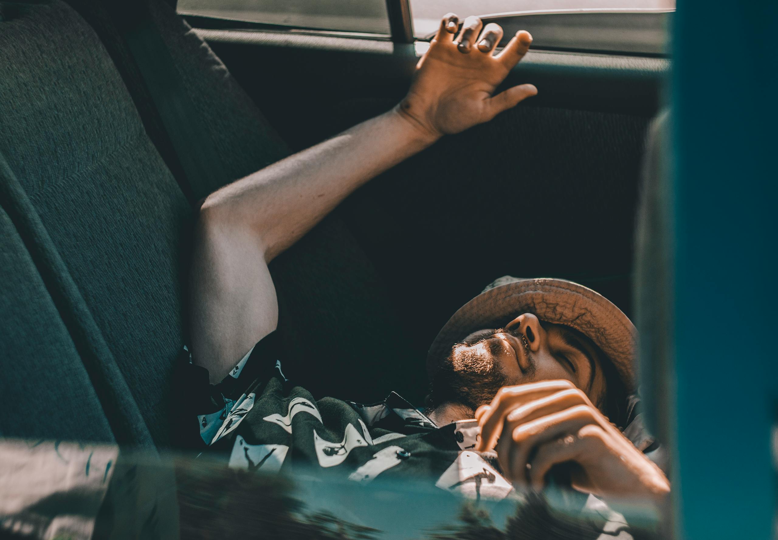 A man with eyes closed rests in the backseat of a car, hand raised.
