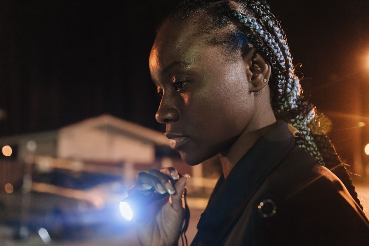 A policewoman with braided hair using a flashlight to investigate an urban crime scene at night.