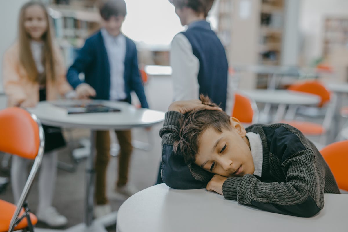 8 Ways In Which Divorce Tears The Family Apart 2 A sad child rests on a classroom desk while peers talk, symbolizing loneliness and school exclusion.