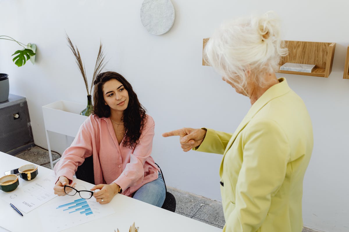 9 Traits Baby Boomers Get Criticized for the Most 1 A senior woman in leadership discussing with a young businesswoman at a modern office desk. boomer Finger-Pointing at Younger Generations