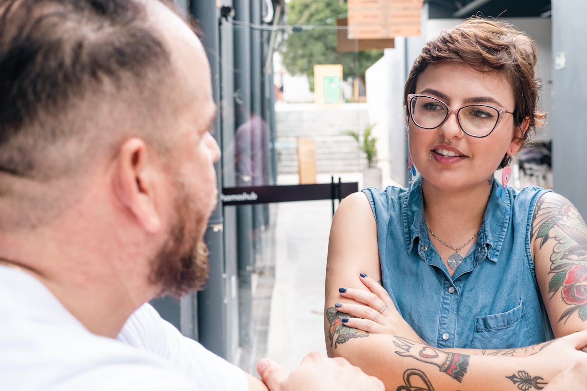 A tattooed woman in a denim shirt and eyeglasses chats with a bearded man outdoors on a sunny day.