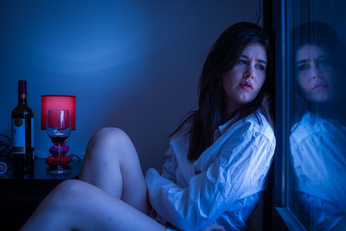 A thoughtful woman in white shirt gazing out a window beside a wine bottle.