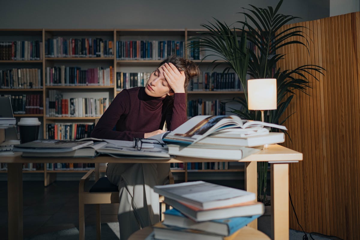 A tired woman surrounded by books, studying in a library, feeling overworked and stressed. 10 Critical Warning Signs You're on the Road to Burnout