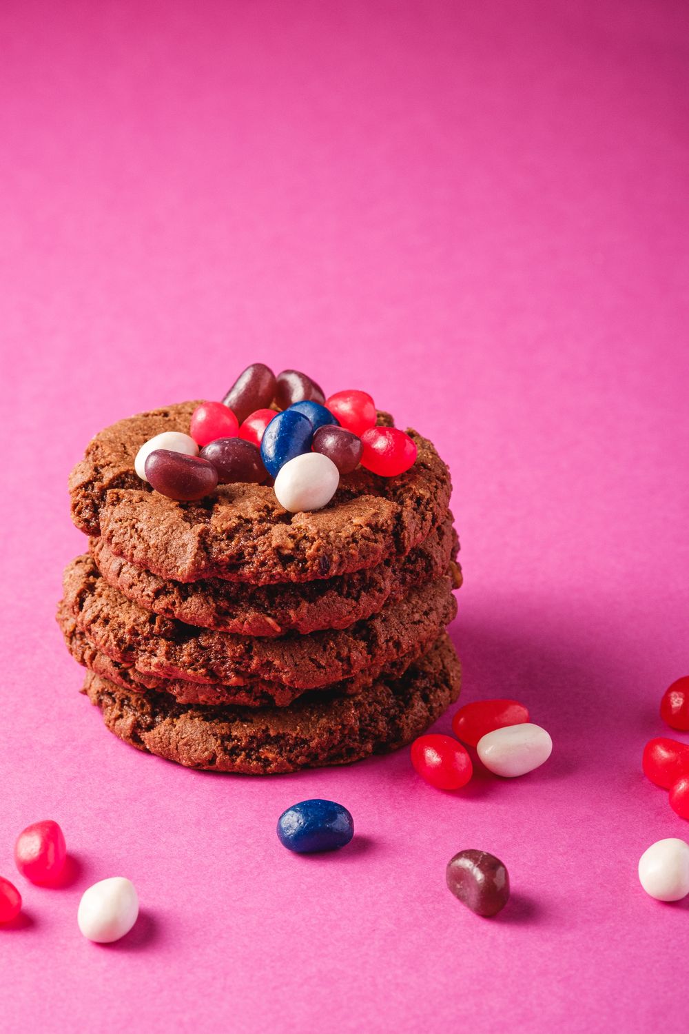 A vertical shot of chocolate cookies stacked with jelly beans on a pink background.