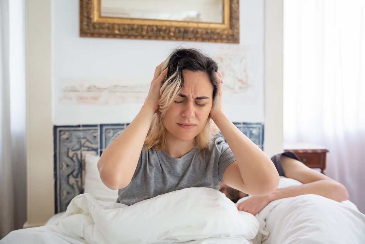 A woman holding her head in discomfort while sitting on a bed in a sunlit room.