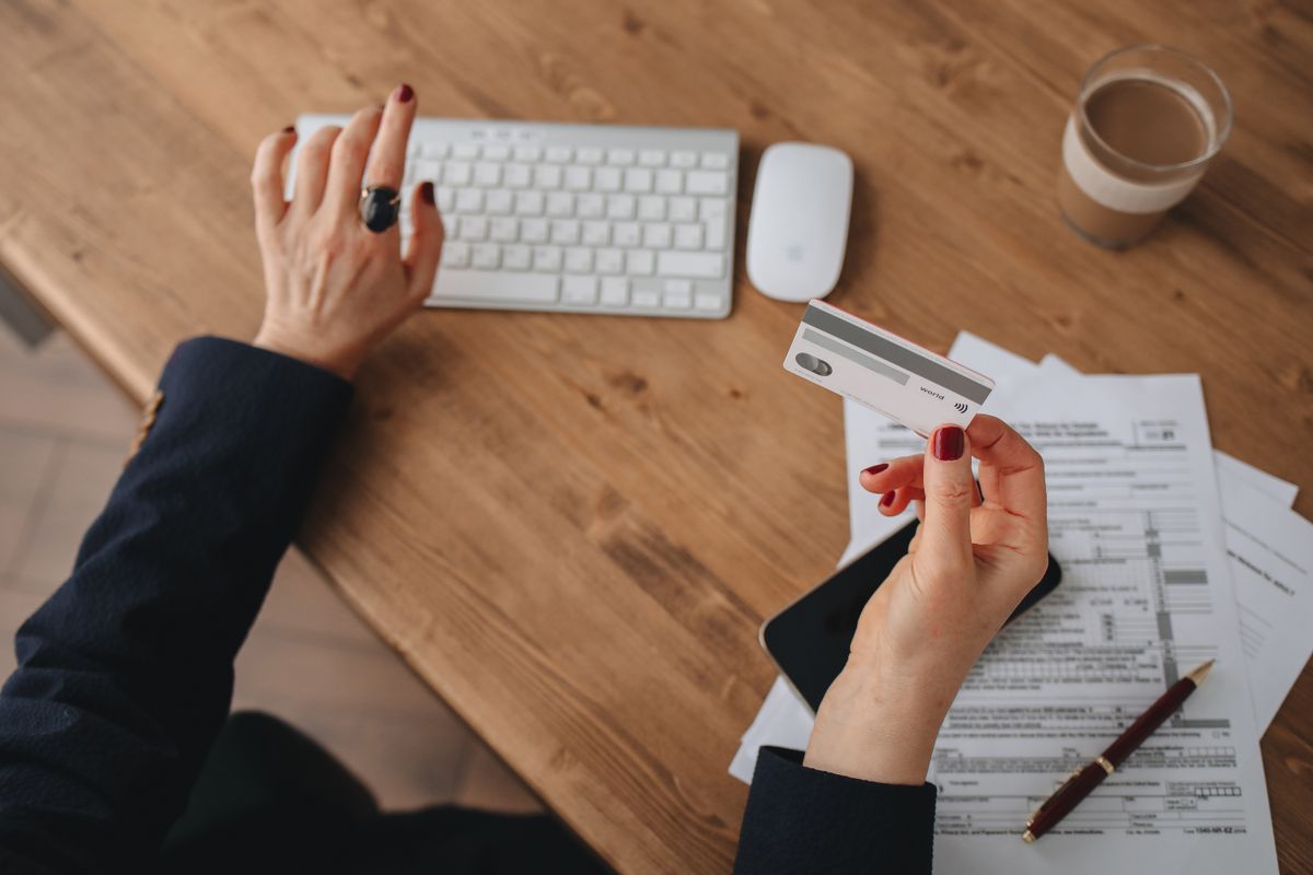 What Happens If You Do Not Pay a Deceased Person’s Credit Card Debt 1 A woman uses an Apple Magic Keyboard while holding a credit card over paperwork on a wooden desk.