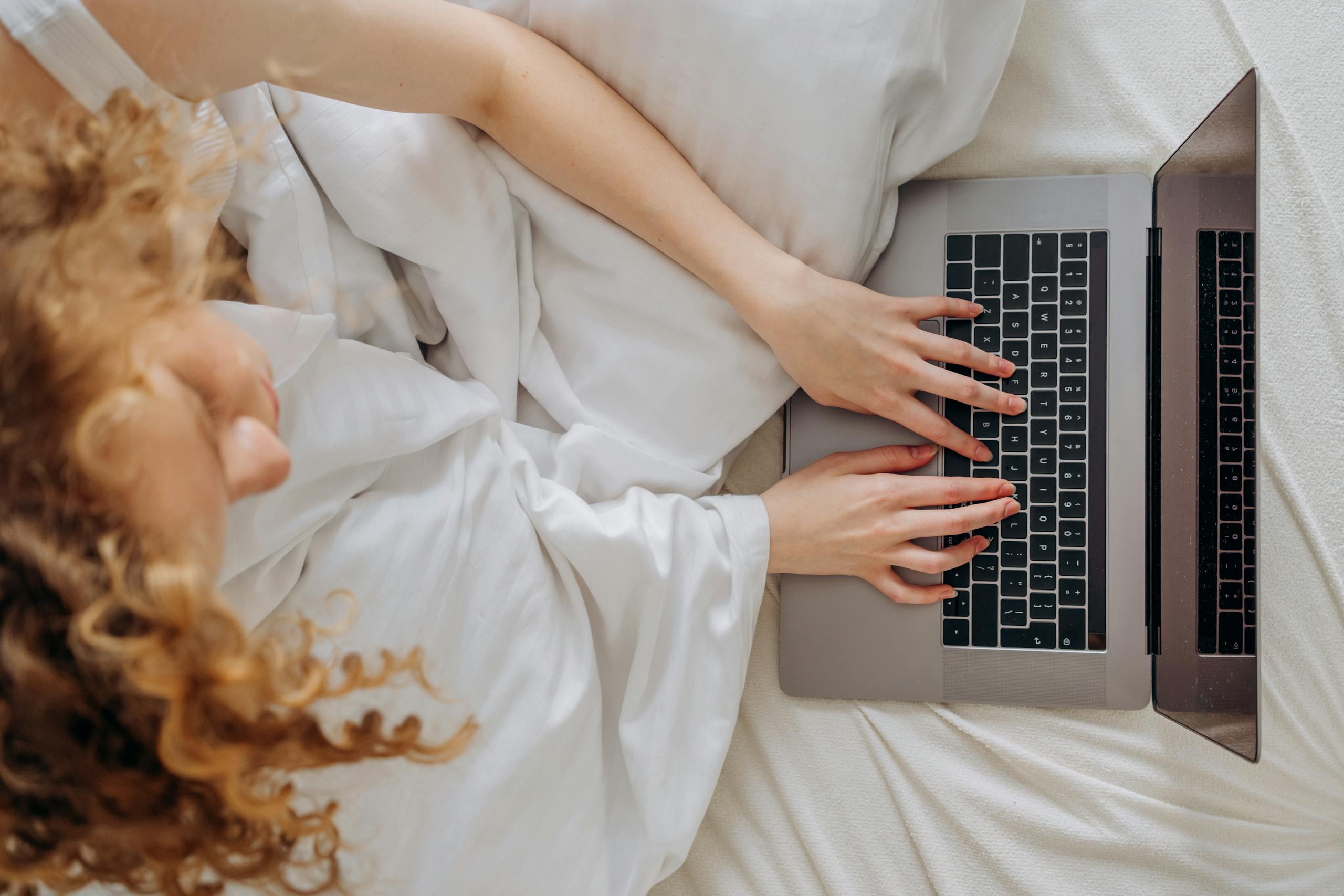 A woman with curly hair working on a laptop while relaxing in bed, photographed from above.