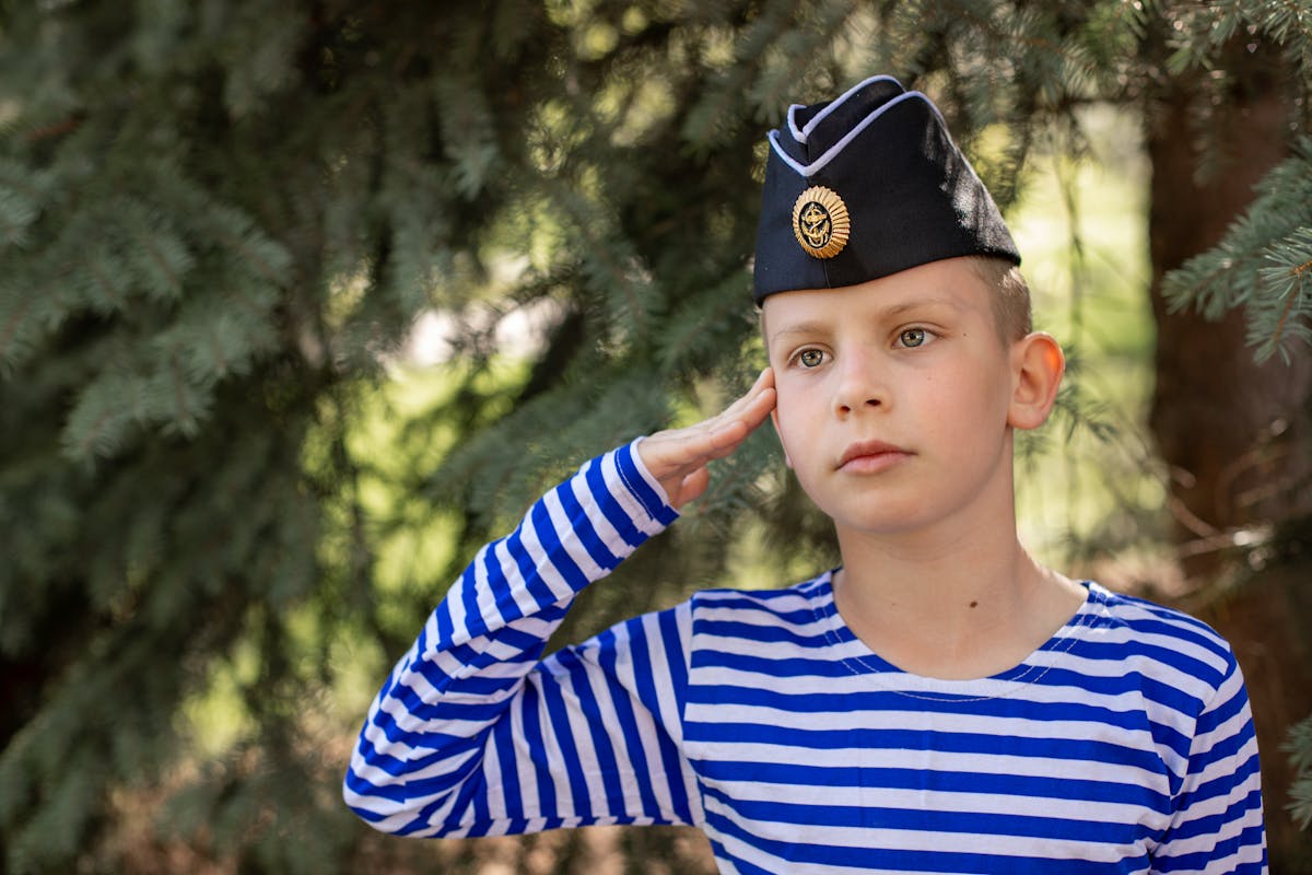 The Military Salute You’ve Been Misreading Your Whole Life 4 A young boy in a striped shirt saluting while wearing a forage cap outdoors.