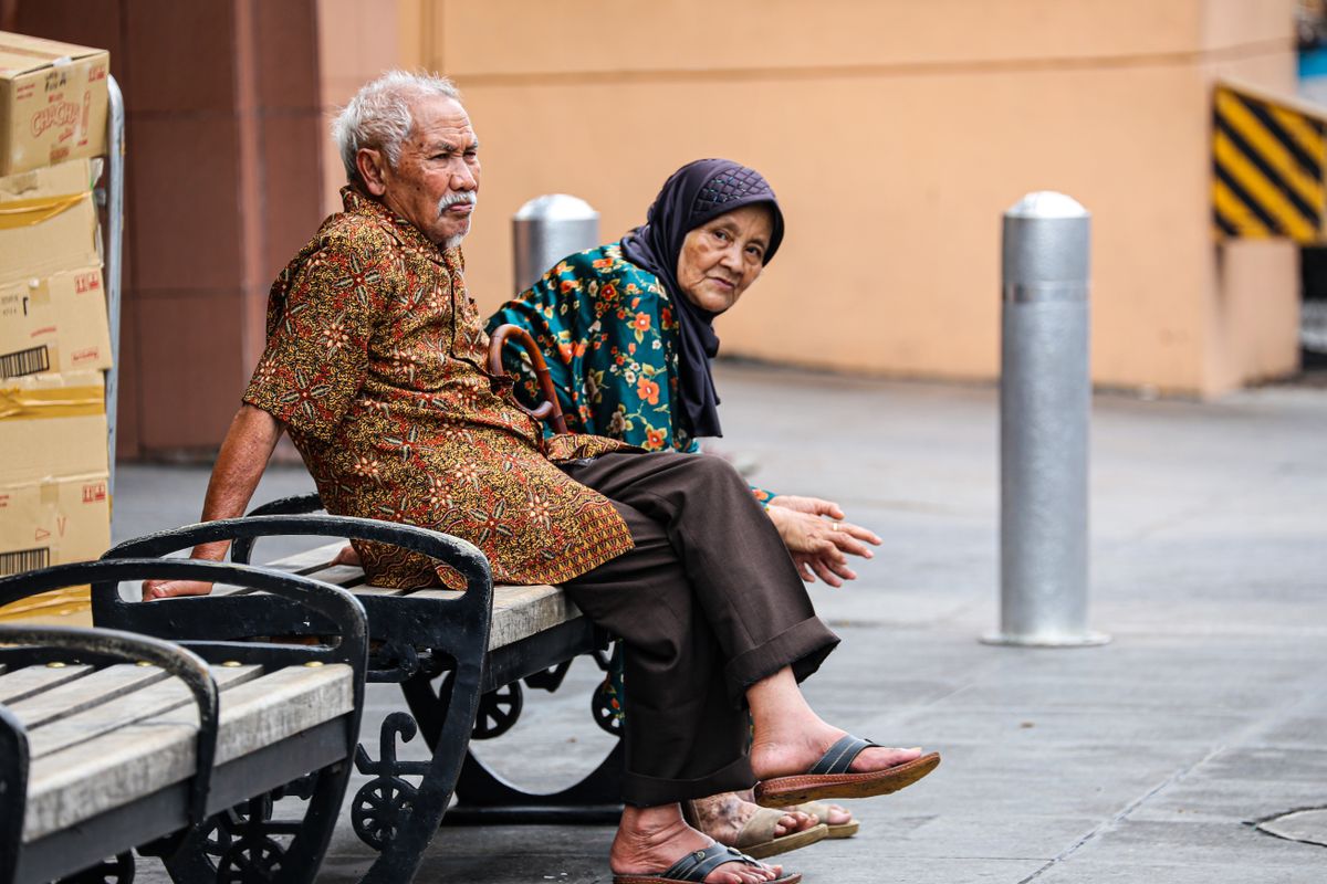An elderly couple in traditional attire sits on a bench in Yogyakarta, Indonesia.