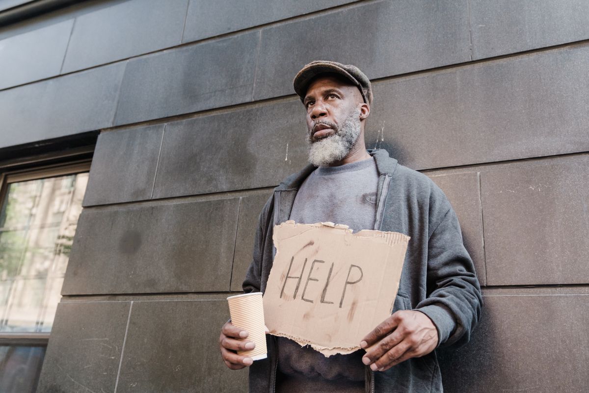 Florida Cities Are Forcing Out the Poor and Elderly Living in RVs in 2026 3 An elderly homeless man holds a cardboard sign asking for help in an urban setting.
