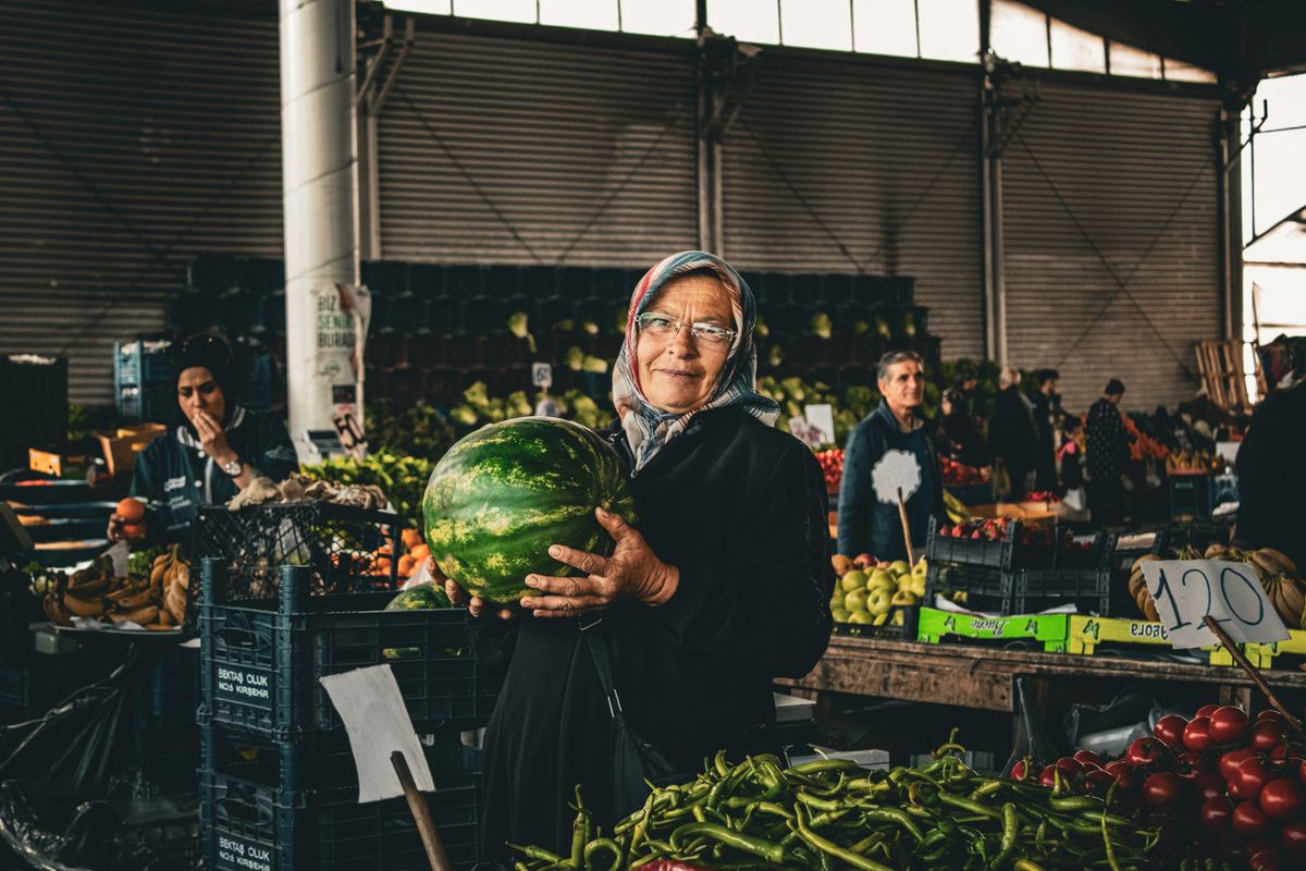 An elderly woman in a headscarf holds a watermelon at an indoor produce market.