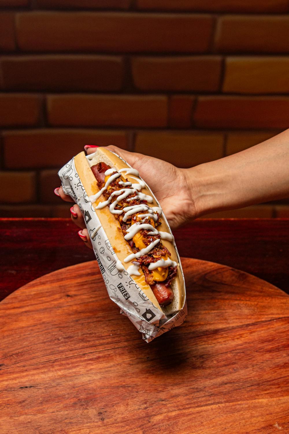 Appetizing hot dog with toppings and sauces held by a hand against a warm background.