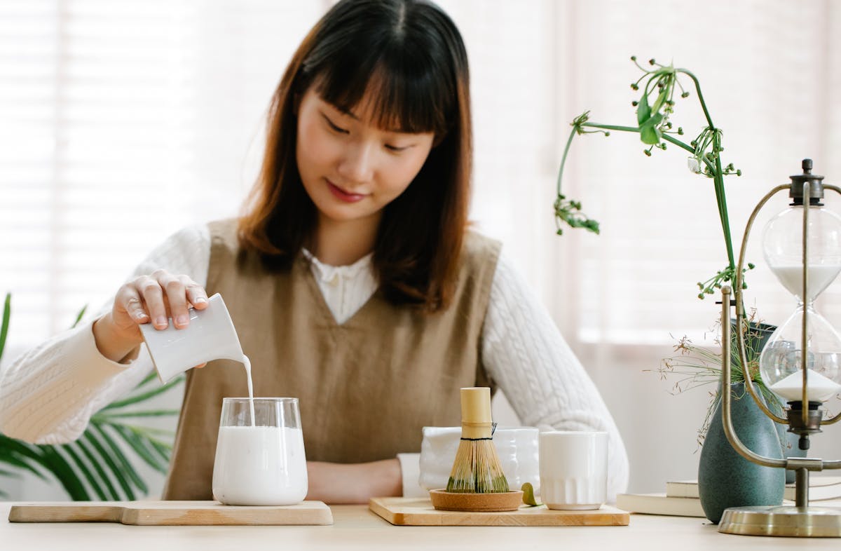 Asian woman pouring milk into a glass in a minimalist indoor setting, with a bamboo whisk and plants.