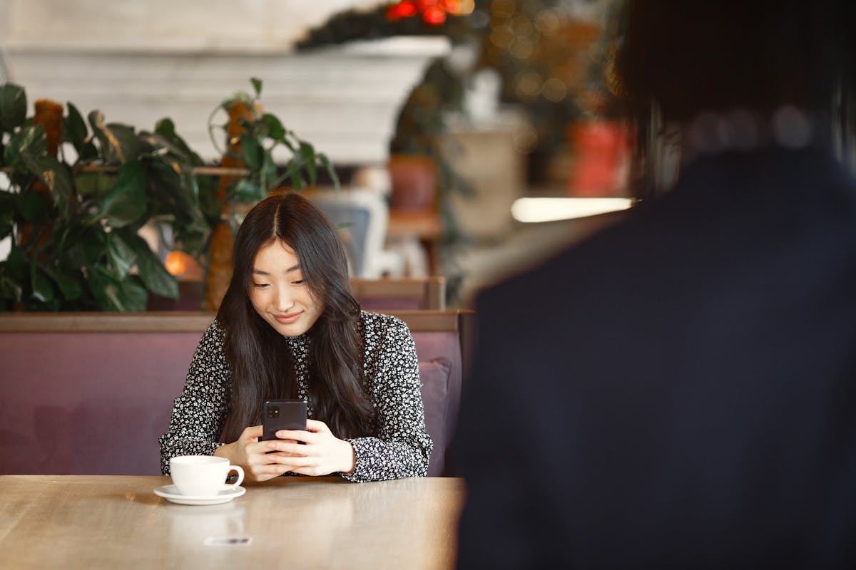 Asian woman with long hair smiling while using smartphone in a cozy cafe, enjoying a cup of coffee.