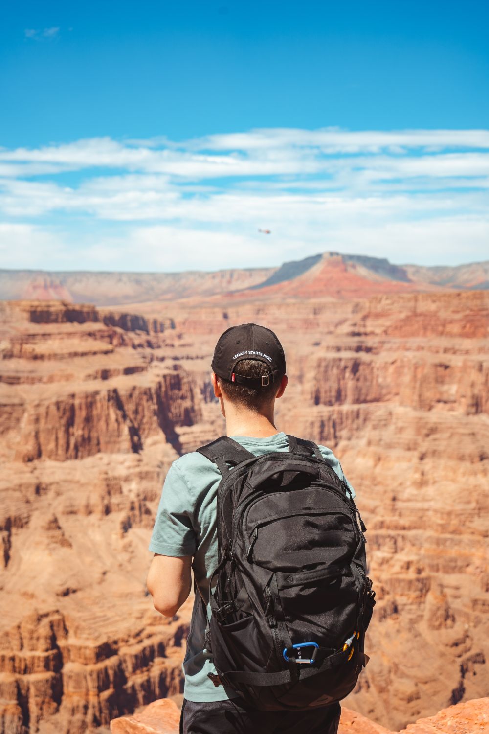 Back view of unrecognizable young male traveler in cap and casual clothes with backpack admiring picturesque scenery of Grand Canyon against cloudy blue sky