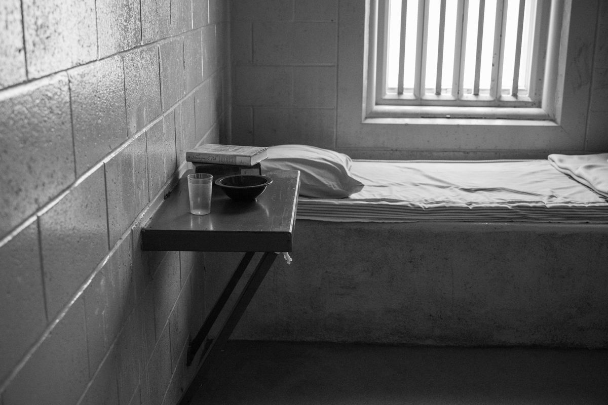 Black and white image of a minimalist prison cell with a bed, table, and window.
