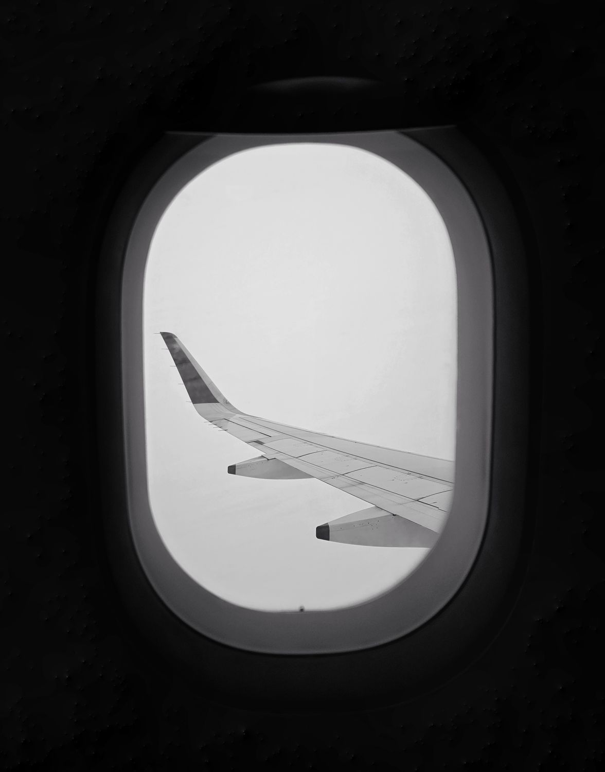 Black and white shot of airplane wing seen through window mid-flight, minimalist aesthetic.