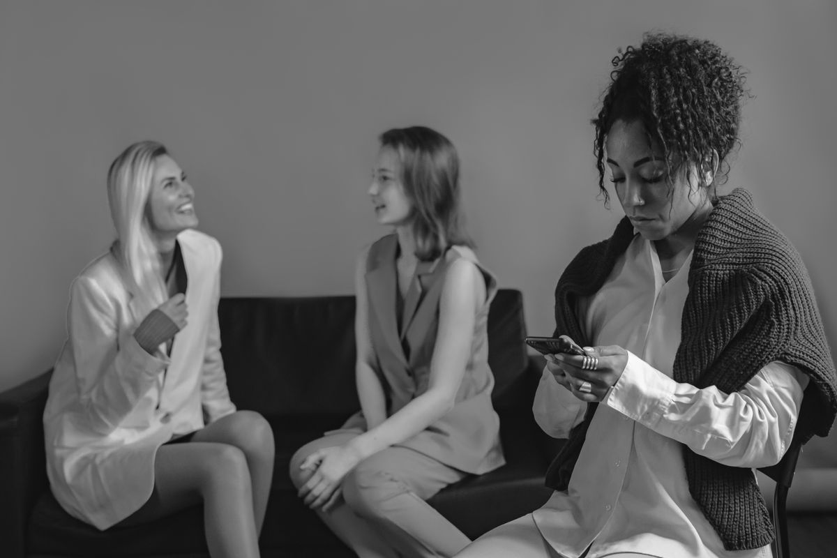 7 Ruthless Signs Someone Is Secretly Sabotaging Your Success 4 Black woman using phone while seated apart from two chatting women on couch.