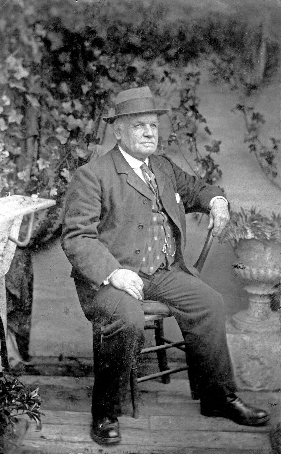 Classic vintage photograph of a senior man seated outdoors wearing a suit and hat.