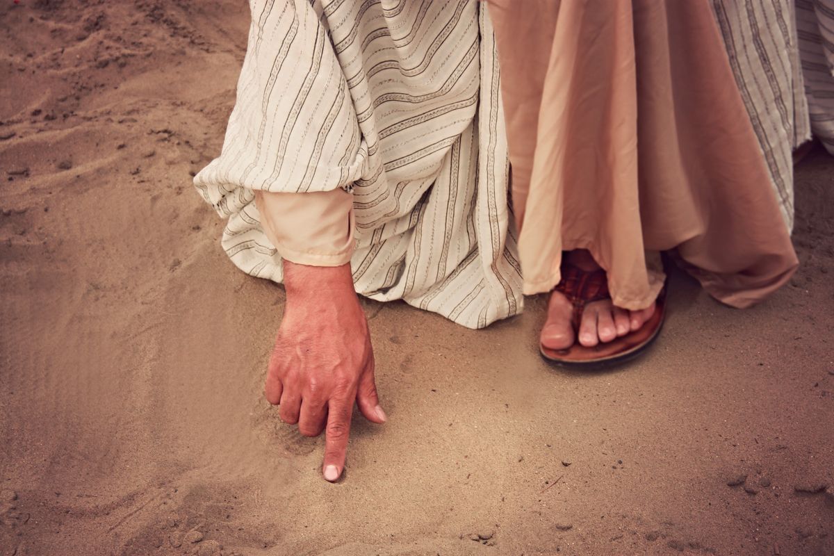 Close-up of a hand pointing on sand, wearing vintage clothing and sandals, emphasizing a cultural or historical theme.