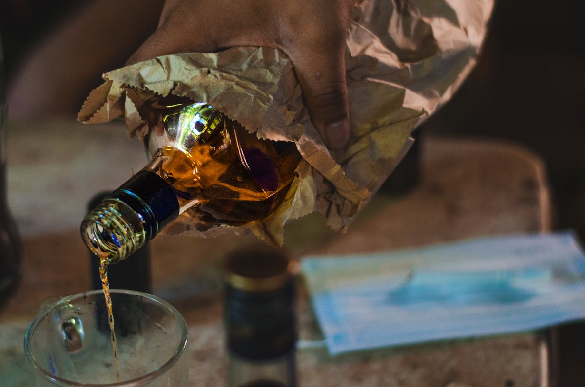 Close-up of a hand pouring whiskey from a paper-wrapped bottle into a glass, indoors. alcolhol
