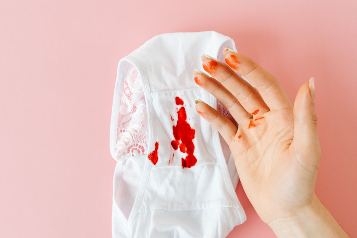 Close-up of a hand with blood stains next to bloodstained underwear on pink background.
