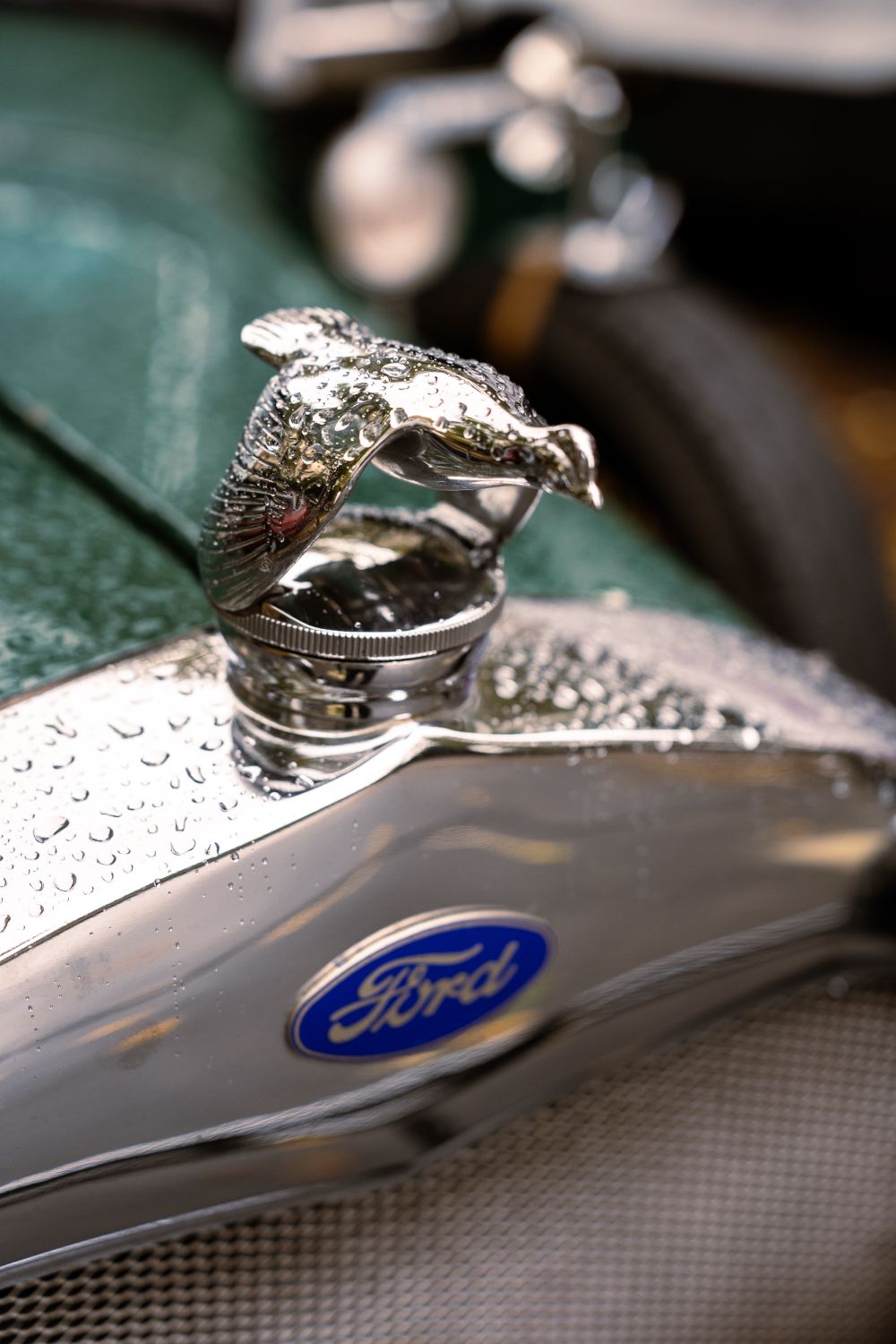 Close-up of a vintage Ford car emblem with a chrome hood ornament and raindrops, showcasing classic automotive design.