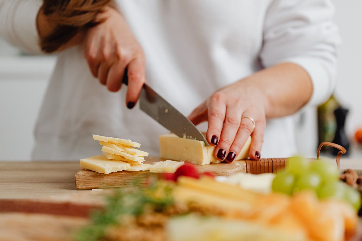 Close-up of hands slicing cheese with fresh fruits in the background, showcasing food preparation. Processed Cheese