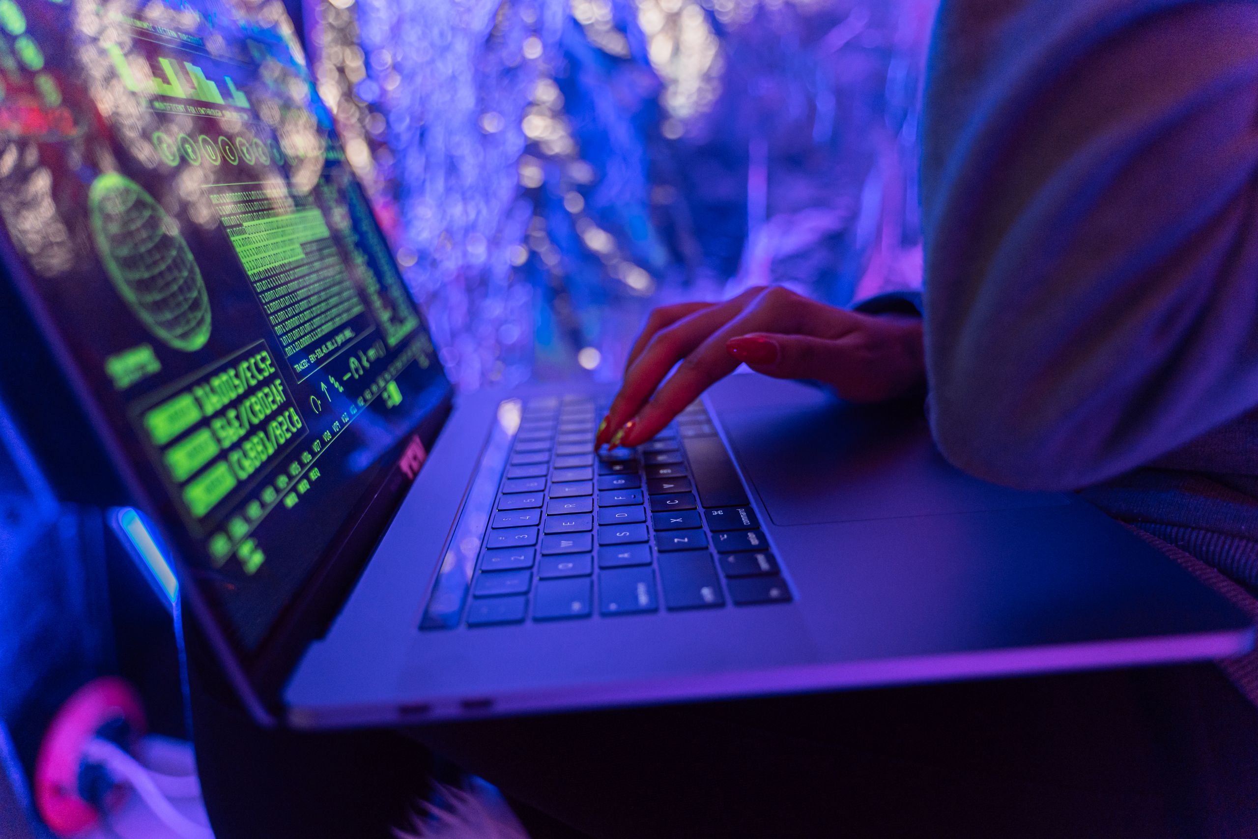 The Hidden Servers Behind a $63 Million Cybercrime Scheme Targeting Americans 1 Close-up of hands typing on a laptop displaying cybersecurity graphics, illuminated by purple light.