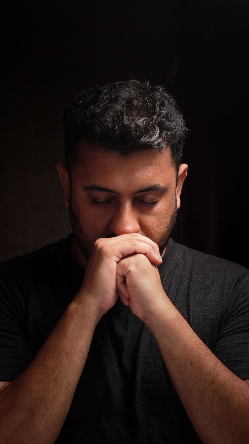 11 Painful Signs God Has Forgiven You, Even If Guilt Still Follows You 3 Close-up portrait of a thoughtful man with hands under nose, eyes closed, in dim lighting.