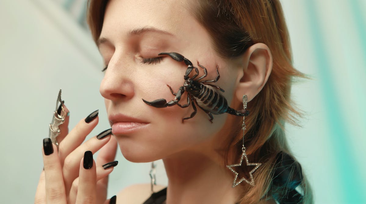 Close-up portrait of a woman with closed eyes, adorned with jewelry and a scorpion on her face.