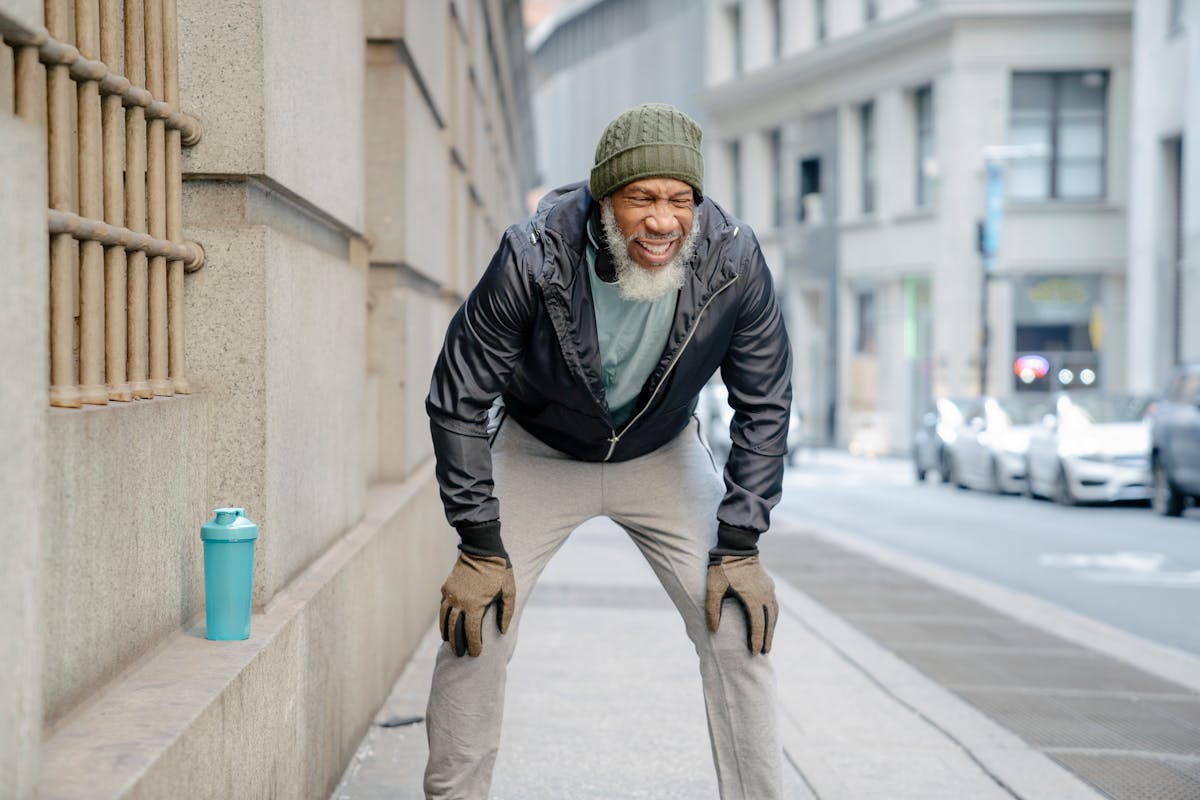 Concentrated bearded mature black man in activewear standing on street leaning forward with hands on knee while resting after hard training